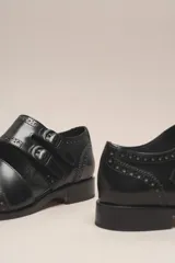 Zapato bajo de cuero negro, estilo monk strap con dos hebillas ajustables sobre el empeine. Presenta detalles de brogue (perforaciones) en la puntera y alrededor del borde superior. Tiene un taco bajo y ancho.