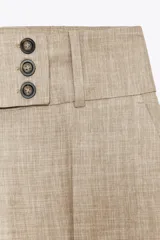 Pantalón de tiro alto color beige, con bolsillos delanteros y falsos bolsillos traseros. Pierna ancha y cierre frontal con ganchos metálicos y botones.