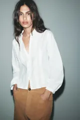 Camisa blanca de cuello redondo y escote pico, con mangas largas abullonadas y detalle de puntillas combinadas a tono en el delantero. Cierre frontal con botones forrados.