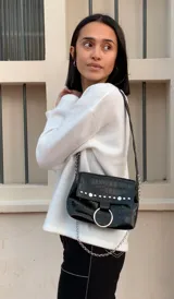 Cartera bandolera negra de ecocuero con textura arrugada, tachas plateadas, cadena decorativa y aro metálico.