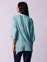 Blusa celeste con estampado de cachemir en blanco, cuello a la base, escote en V y abertura central con botones. Manga 3/4 regulable con presilla y botón, y terminación de ruedo asimétrico.