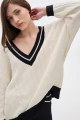 Sweater tejido de punto color crema, con cuello en V con borde negro y doble línea blanca, mangas largas con puños negros, diseño con trenzas en el frente y corte oversize.