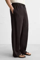 Pantalón relaxed fit color marrón oscuro confeccionado en tejido con mezcla de algodón y lino. Cintura elástica, bolsillos laterales y bolsillo trasero de plastrón. Perneras con pliegues frontales. Cierre en delantero con cremallera y botón.