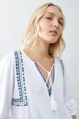Remera blanca de estilo boho con bordados en el escote y mangas largas. Presenta escote en V con lazos y borlas. Corte recto y holgado.