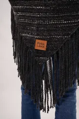 Ruana negra tejida en telar manual con lana merino. Presenta diseño con líneas horizontales en blanco y flecos largos en el ruedo. Incluye bolsillos de cuero natural vacuno.