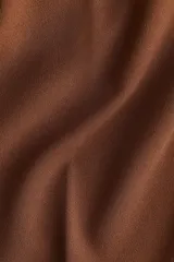 Pantalón tobillero color marrón, de punto pesado, tiro alto, con pliegues delanteros y traseros, elástico en la parte posterior de la pretina, cierre con botón y corchete ocultos, y bajos levemente acampanados.