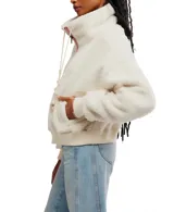 Campera cropped de faux-sherpa color blanco, con cierre frontal de cremallera, cordones trenzados con pompones, cuello alto y bolsillos laterales.