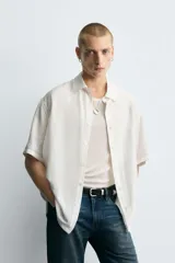Camisa blanca de manga corta con cuello solapa y textura jacquard irregular. Corte relajado (relaxed fit) con aberturas laterales y cierre frontal de botonadura.