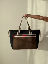 Bolso tipo tote de neopreno color negro y marrón, con cierre superior, bolsillo frontal marrón con logo rojo, bolsillo interno y externo con cierre, doble asa de mano marrón con detalles blancos y opción de correa larga.