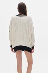 Sweater tejido de punto color crema, con cuello en V con borde negro y doble línea blanca, mangas largas con puños negros, diseño con trenzas en el frente y corte oversize.