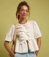 Blusa blanca de tricolina con cuello redondo, manga corta abullonada, cierre frontal con lazos y volado en la cintura.