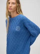 Sweater color crema con tejido en ochos, cuello redondo, puños acanalados con doble raya azul y tajos a los costados.