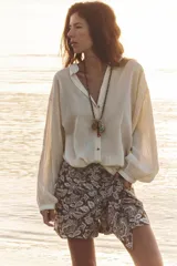 Blusa color crema de tejido arrugado con hilo metalizado, cuello redondo con escote en pico, manga larga abullonada y nudo delantero. Cierre frontal con botones con flores bordadas.