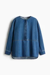 Blusa azul de denim con cuello redondo, tapeta con botones ocultos y mangas largas con puños abotonados.