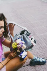 Llavero con tarjeta identificadora transparente con borde violeta, dije de nube blanca con cara sonriente, borla de hilos negros, mosquetón y argolla violeta, cinta de soga trenzada con diseño geométrico en amarillo, violeta y blanco.
