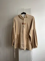 Canguro beige de plush suave con capucha, bolsillo frontal, manga globo y puños fruncidos. Tiene tajo a los lados con tiras ajustables.