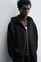 Sudadera negra con capucha, corte boxy, cierre frontal con cremallera, bolsillos laterales y puños elásticos.