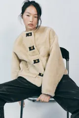 Chaqueta corta de cuello redondo y manga larga en tejido de pelo sintético color negro. Cierre frontal con botones metálicos dorados y bolsillos de plastrón. Colección Zara Woman.