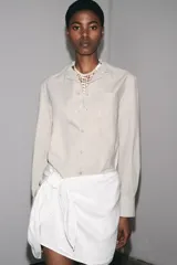 Camisa de manga larga color crudo, confeccionada en hilatura de algodón. Tiene cuello solapa, bolsillo de plastrón en el pecho, bajo redondeado y cierre frontal con botones.