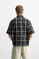 Camisa de hombre relaxed fit con estampado de cuadros blancos sobre fondo negro, cuello solapa, manga corta y cierre frontal con botones.