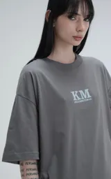 Remera de algodón premium color gris concreto, con corte oversize, hombros caídos y cuello redondo. Presenta un estampado frontal con las siglas "KM" y el texto "#KILOMBITOMAL" debajo en color celeste pálido.
