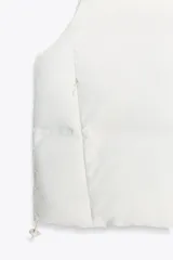 Chaleco blanco acolchado con cuello alto y cierre frontal con cremallera. Tiene bolsillos delanteros con cierre de cremallera oculta y bajo ajustable con cordones elásticos laterales.