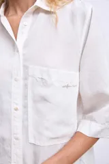 Camisa blanca de manga tres cuartos, con cuello camisero y corte holgado. Presenta un bolsillo de parche en el pecho izquierdo.