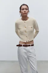 Chaqueta color crema de tejido de punto con cuello redondo y manga larga. Tiene bolsillos delanteros de plastrón y cierre frontal con botones dorados.