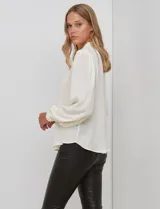 Blusa de gasa color crema o blanco roto, de manga larga con puños ajustados por elástico y botones. Presenta un lazo largo en el cuello que cae sobre el frente.