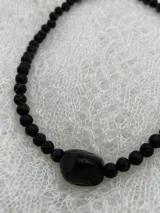 Collar corto de cuentas redondas de piedra volcánica negra, con cuenta central irregular de piedra negra pulida y cierre de gancho dorado.