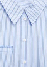 Camisa corta celeste a rayas verticales, de corte holgado, con cuello camisero, mangas largas con puños abotonados, bolsillo de parche en el pecho y cierre frontal con botones. Detalle de costuras a contratono.