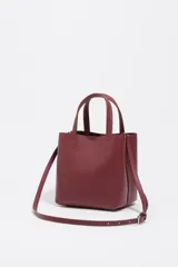 Bolso pequeño tipo tote o cubo de piel vacuna texturizada color negro, con asas cortas superiores y correa larga ajustable para llevar al hombro o cruzado. Presenta el logo metálico de Bimba y Lola en dorado en el frente.