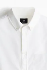 Camisa blanca de algodón Oxford con cuello americano, tapeta clásica, bolsillo superior abierto y canesú en la espalda. Mangas largas con puños abotonados y bajo levemente redondeado. Corte estándar.