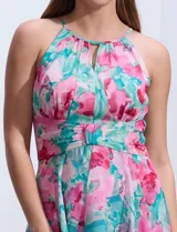 Vestido largo de gasa estampada con flores rosas y turquesas sobre fondo blanco. Tiene escote halter con ojal, cintura fruncida y falda con sobrecapa.