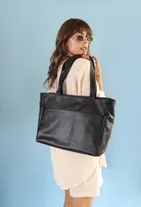 Cartera tipo tote de cuero graneado color negro, con textura uniforme y suave al tacto. Tiene manijas de 55 cm, bolsillo frontal con broche imantado, bolsillo trasero con cierre metálico, cierre principal metálico, forro interior y tres bolsillos internos (uno con cierre, uno plaqué y uno tipo sobre con solapa y botón).