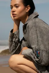 Vestido camisero de denim gris con cuello camisero, cierre frontal con botones, mangas largas con puños abotonados, bolsillos laterales y apliques bordados en el pecho y la manga. Corte holgado y largo a la rodilla.