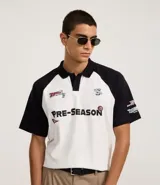 Remera boxy de algodón blanca con mangas raglán negras, cuello polo negro y estampa frontal con la leyenda 'Pre-Season' y otros logos.