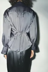 Camisa gris de satén con cuello solapa, escote en pico, mangas largas abullonadas y cierre frontal con botones metálicos.