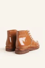 Bota de cuero vacuno con acabado charol, color marrón oscuro. Presenta un diseño de caña corta con cordones frontales y costuras decorativas en la puntera. La suela es de goma tipo crepe.