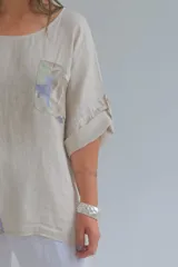 Blusa beige de lino con mangas 3/4 y apliques de tela estampada con motivos florales en bolsillo y parte inferior.