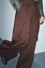 Pantalón ancho de tiro medio color marrón oscuro, confeccionado en tejido con lana. Presenta pinzas delanteras, cinturón ajustable con hebilla metálica y bolsillos cargo de plastrón en las piernas.