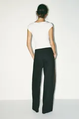 Pantalón negro con finas rayas verticales blancas, de tiro medio y corte ancho. Presenta bolsillos delanteros y traseros, cintura doble tipo bóxer con botones y cierre frontal con cremallera y ganchos metálicos. Detalle de franja marrón en los laterales.