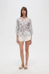 Blusa blanca de algodón con bordado broderie inglés y estampado floral en tonos azules y marrones. Posee cuello en V con abertura y botones frontales, y mangas tres cuartos.