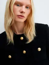 Chaqueta negra de tweed con cuello redondo, cierre frontal con botones dorados, bolsillos de parche y flecos en el ruedo.