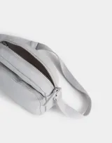 Bandolera gris claro de tela deportiva con bolsillo frontal, correa larga regulable, acceso con cierre superior y tag de goma.