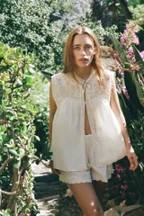 Blusa sin mangas color blanco con cuello alto, bordados y puntillas combinadas a tono, y apliques de abalorios. Cierre frontal con abertura, lazada y botón.