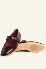 Zapato tipo Mary Jane de charol color borgoña, con punta redondeada, tira sobre el empeine con hebilla y suela de cuero.