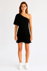 Minivestido negro asimétrico con un hombro descubierto y manga abullonada. Presenta frunces en la falda y volados en el ruedo.