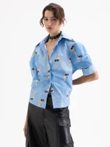 Camisa de poplin satinado color celeste con estampado de cerezas y rayos amarillos. Presenta mangas tipo bombé con puño y cierre frontal con botones ocultos.