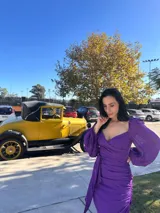 Vestido violeta con escote cuadrado, mangas largas abullonadas, corte ajustado y detalle drapeado con lazo en la cintura.